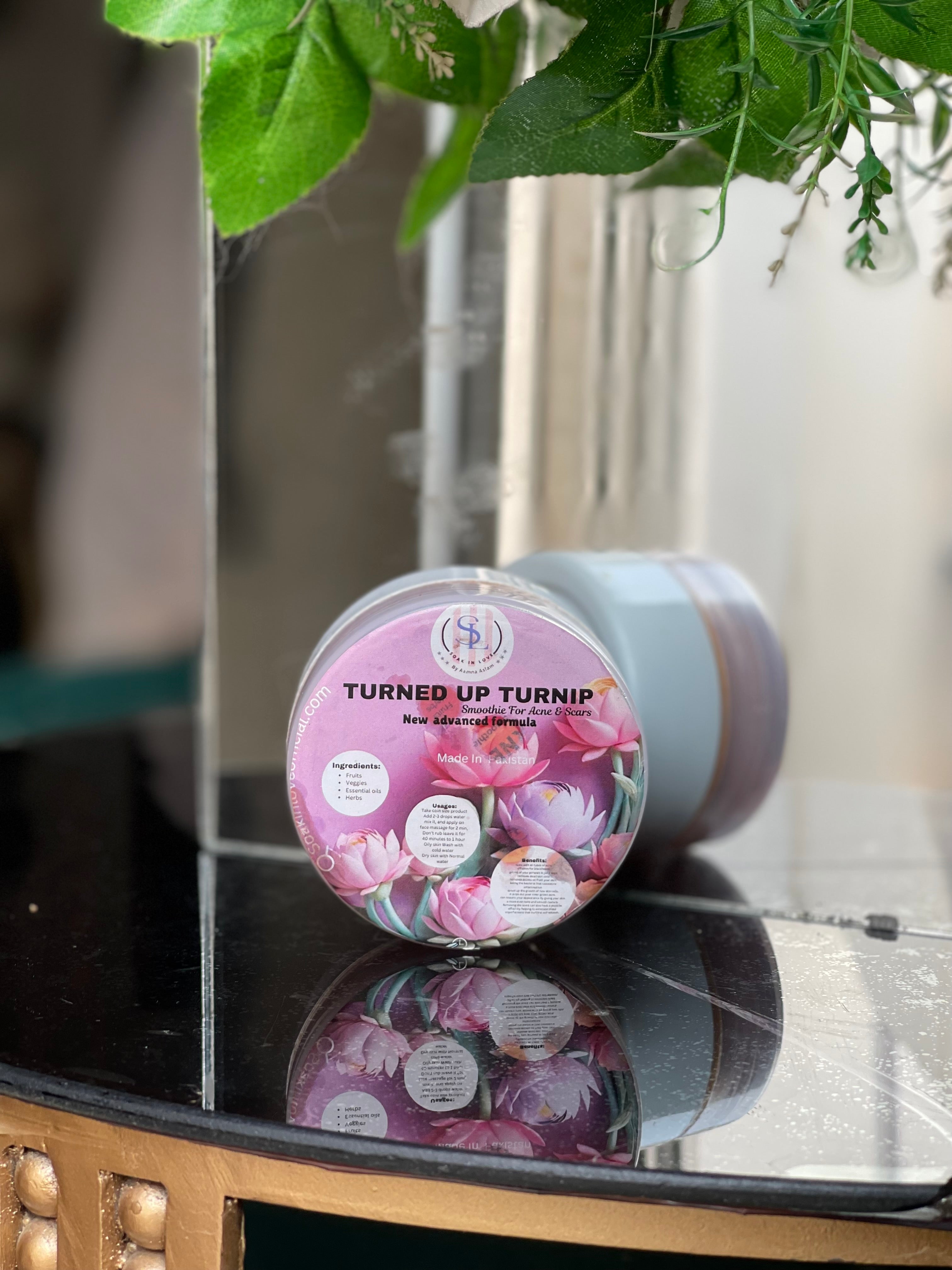 A lavender Turned Up Turnip jar by Soak in Love rests on a reflective surface adorned with green leaves above. The label, showcasing pink floral designs, emphasizes targeted skin care benefits, specifically for acne treatment and minimizing acne scars.