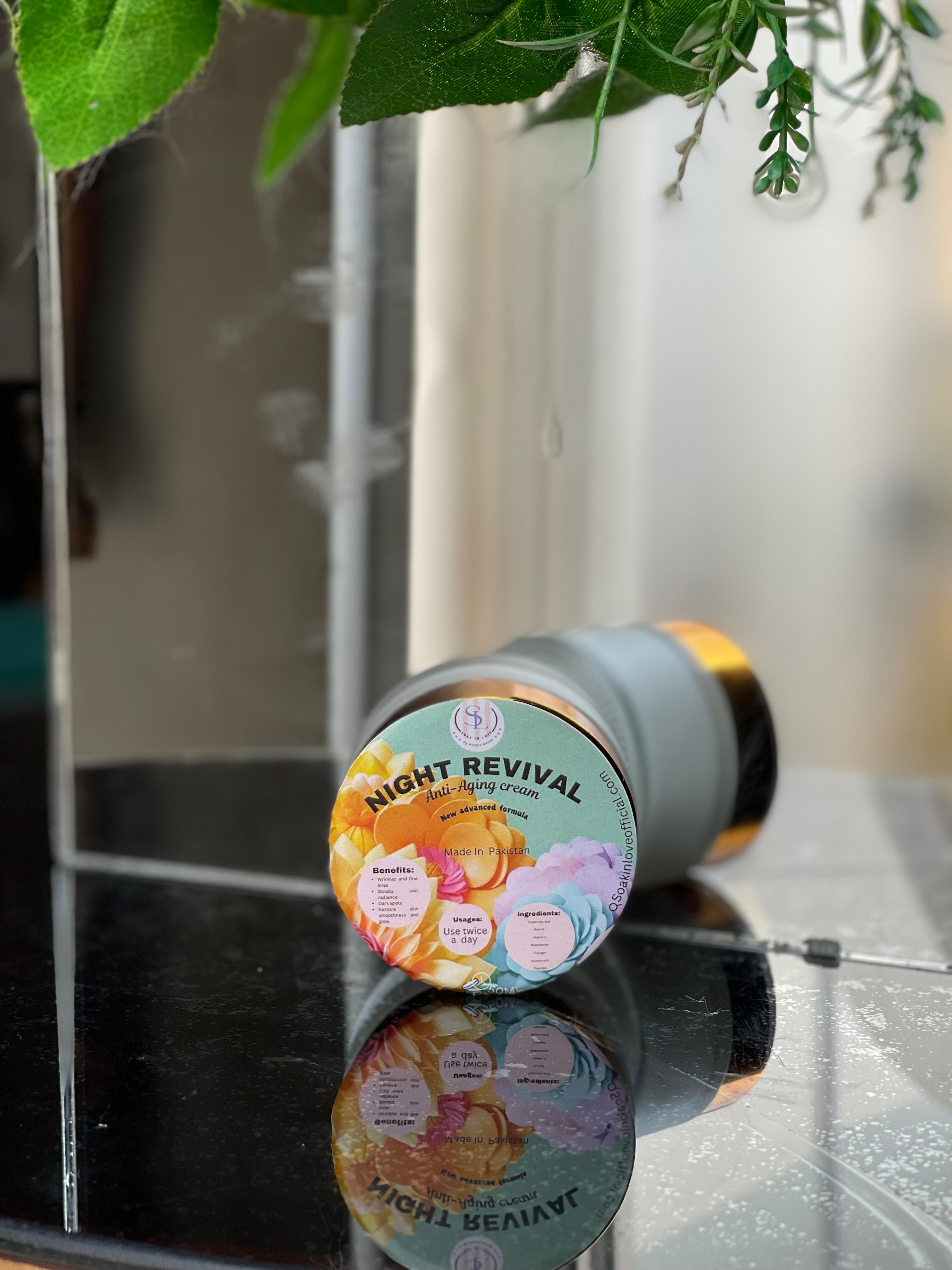 A jar of Revival Night Cream from My Store sits on a reflective black surface, with green leaves cascading in the backdrop. Its label is adorned with colorful flowers and text highlighting its anti-aging benefits, such as effective moisturizing and firming.