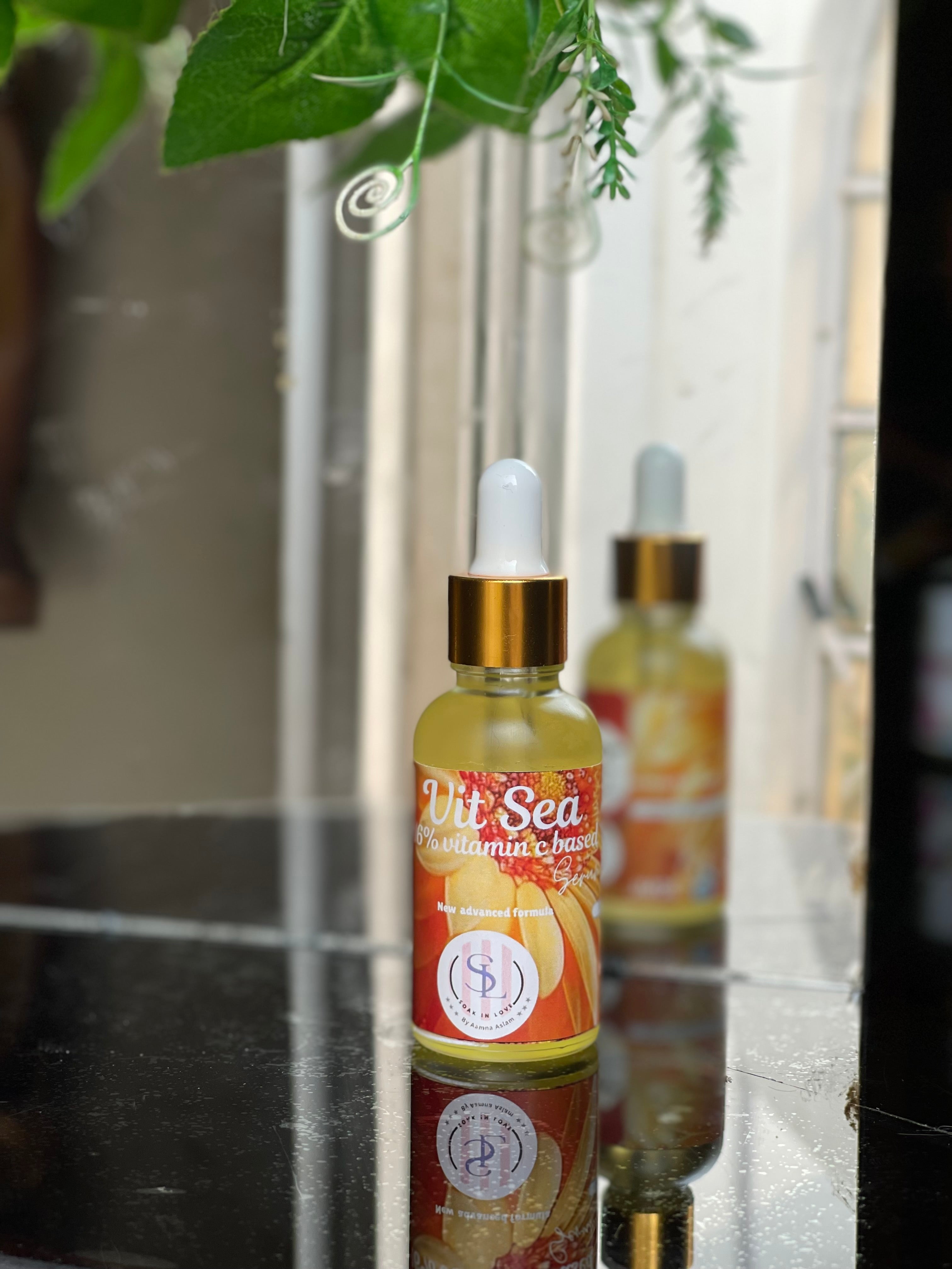 A bottle of Vit Sea Serum from Soak in Love, featuring a dropper cap, sits on a reflective surface with vivid orange accents on its label. Highlighting its antioxidant skincare benefits, a leafy plant hangs overhead while a window is in view, as the mirrored bottle assures radiant, anti-aging effects.