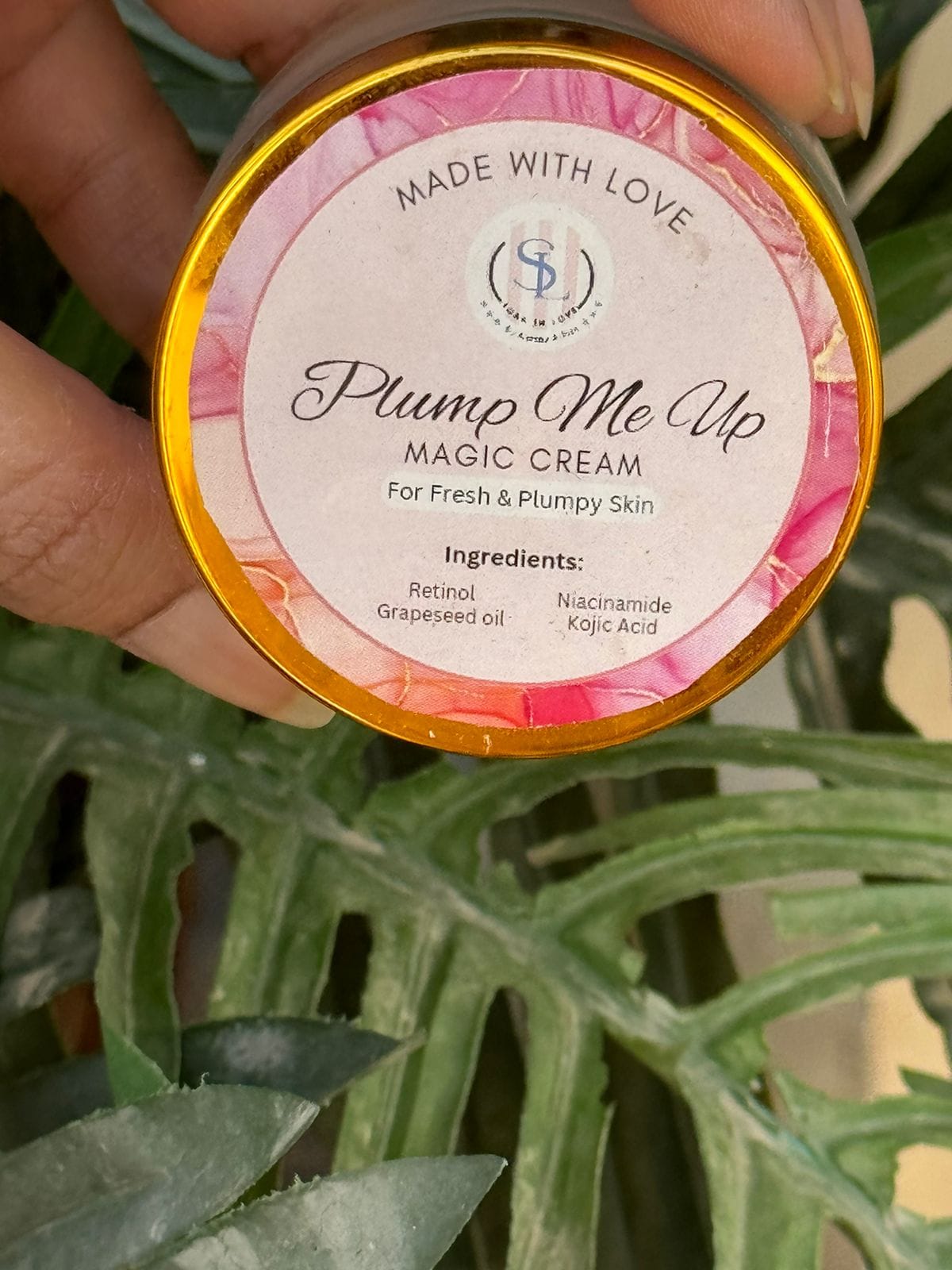 A hand holds a jar of Plump Me Up (Magic Cream) from Soak in Love, accentuated by its pink and gold label. The text says "Made with Love" and highlights natural ingredients such as retinol, grapeseed oil, niacinamide, and kojic acid. Set against a leafy backdrop, this cream also boasts the Radiance Lift Pearl Complex for an added glow.