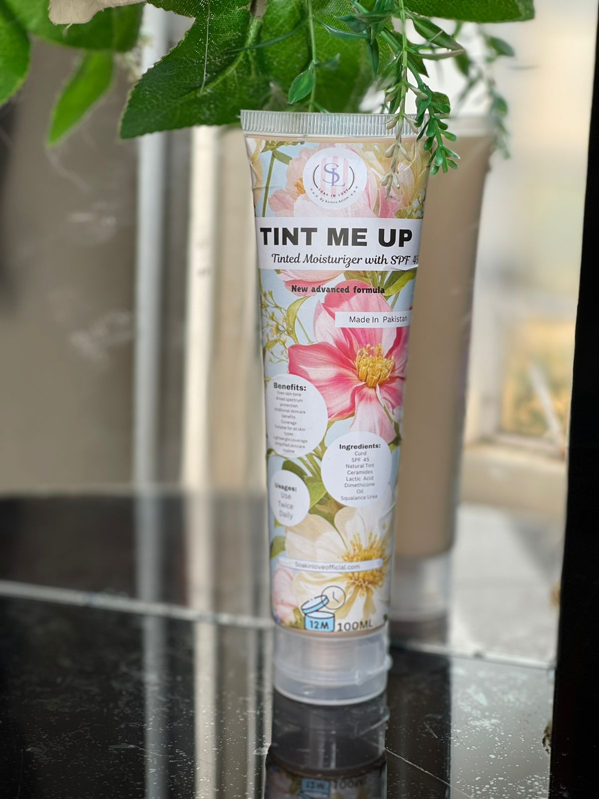 A tube of My Store's Tint Me Up tinted moisturizer, boasting hydrating skin benefits and SPF 30 for sun protection, is elegantly displayed on a reflective surface. Its packaging, featuring floral designs and ingredient highlights, stands gracefully with a green plant in the background.