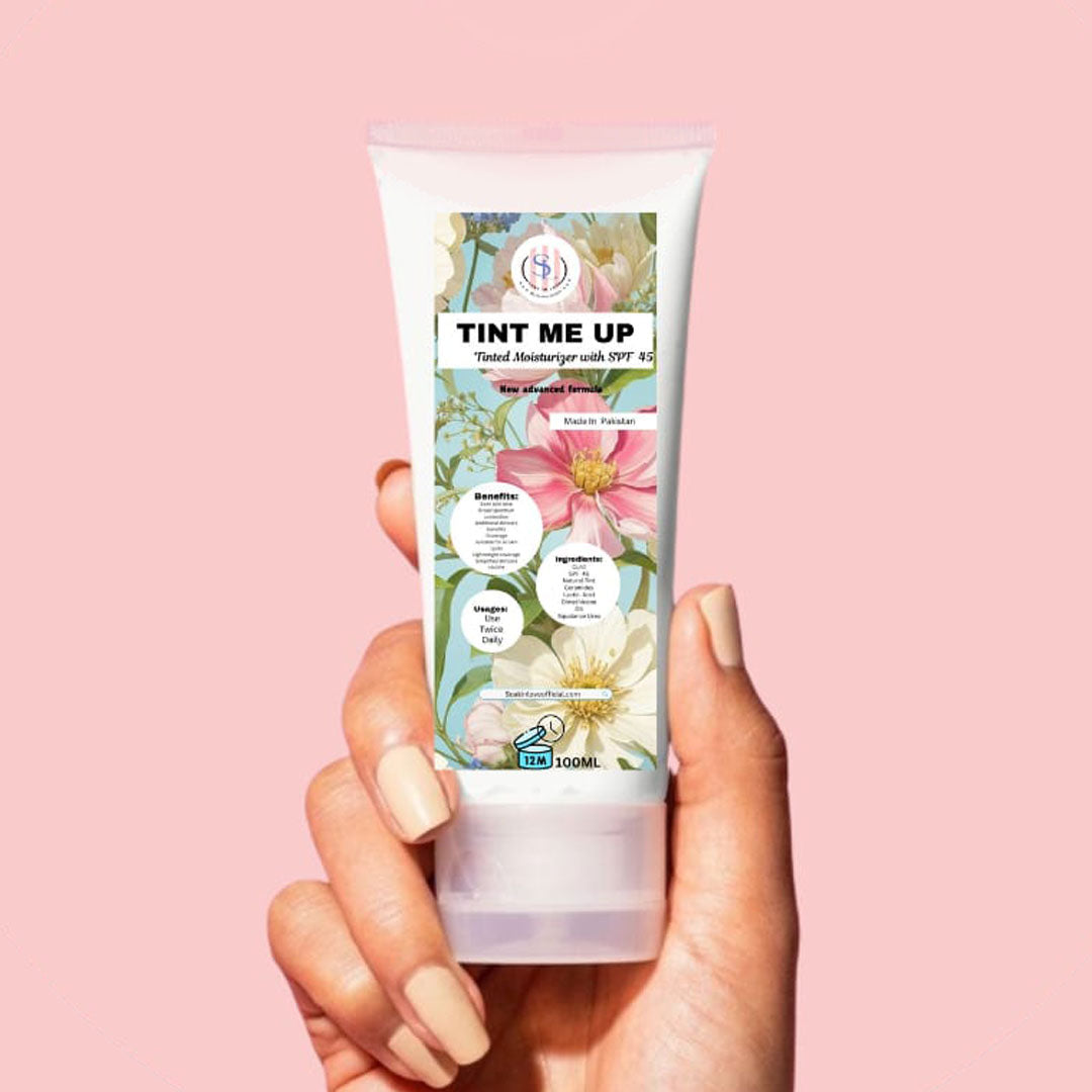 A close-up of a hand holding a My Store's Tint Me Up tube against a pink background. The label is adorned with floral designs and emphasizes SPF 45 sun protection, a lightweight formula, and antioxidant benefits. This 100 ml BB cream is perfect for hydrating skin.